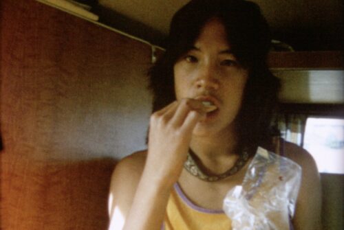 An Asian American teenage boy with shoulder-length hair looks into camera, wearing a yellow tank top and eating a snack out of a plastic bag.