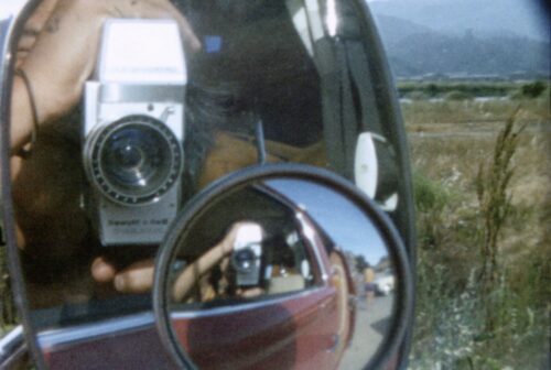A car passenger films directly into the side-view mirror, where their hands and camera can be seen in both the main mirror, and a smaller mirror within– providing a warped 