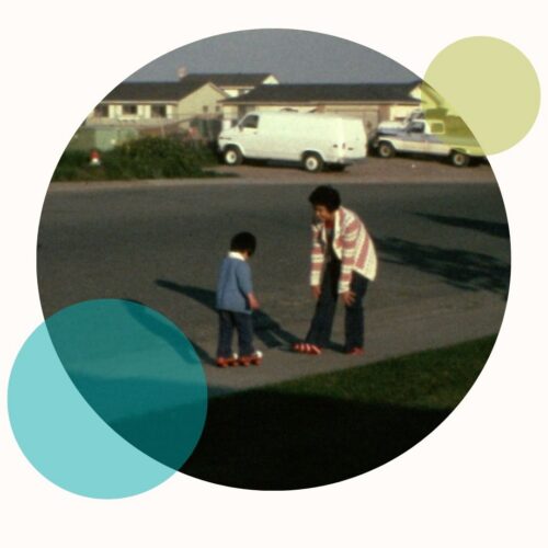 A young Chiense-American boy wearing roller skates stands on a suburban sidewalk, making his way to his mother, who faces him, also wearing roller skates.