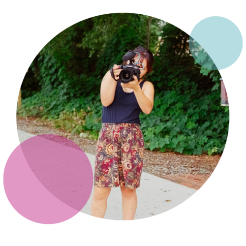A picture with a woman in a black tank top and flowery pants looking through the camera and pointing it towards the viewer.