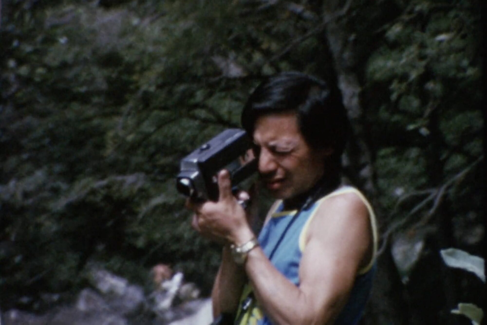 An Asian American man shoots film, looking into the camera with one eye.