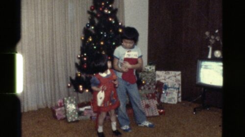 An older brother and younger sister (both Chinese American) look at each other, standing in the living room in front of a colorfully-lit Christmas tree and wrapped gifts. The boy holds a stocking in his hands.