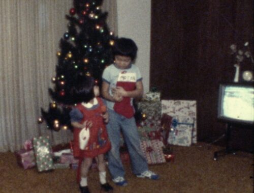 An older brother and younger sister (both Chinese American) look at each other, standing in the living room in front of a colorfully-lit Christmas tree and wrapped gifts. The boy holds a stocking in his hands.