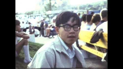 A Chinese American man with glasses looks into camera, with groups of people behind him sitting on a grassy hill and a nearby bench. The young man in-image is CAAM's past Executive Director: Stephen Gong.