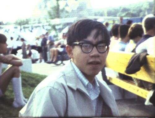 A Chinese American man with glasses looks into camera, with groups of people behind him sitting on a grassy hill and a nearby bench. The young man in-image is CAAM's past Executive Director: Stephen Gong.