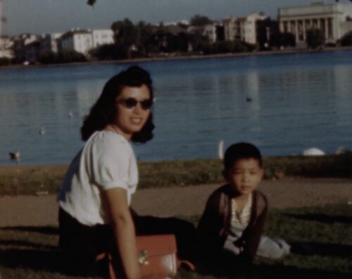 A smiling Chinese American mother with sunglasses sits besides her son. Both look directly at camera, with Oakland's Lake Merritt behind them in the background.