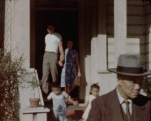 Led by their grandfather, a very young boy and girl hold hands and make their way out of their front door, walking down a Victorian-style staircase. A man and women behind them, likely parents, follow out the door. All members shown are Chinese American.