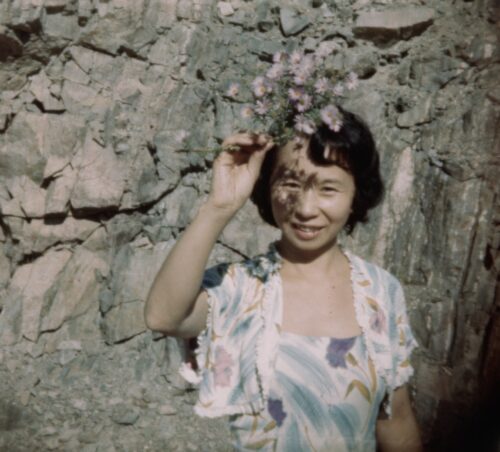 A portrait-style image of a Japanese American woman, holding a bouquet of wild flowers above her head, with scattered shadow overlaying half of her face. She wears a colorful dress and shawl and stands in front of a natural rock wall.