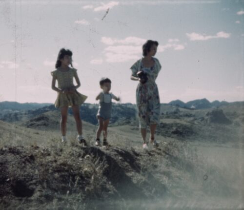 Japanese American members of the Sasaki family (mother, daughter, and young son) stand upon a dry, grassy hillside, gazing to the right of frame to take in the scenery. They are backed by rolling hills and a sunny sky with clouds.